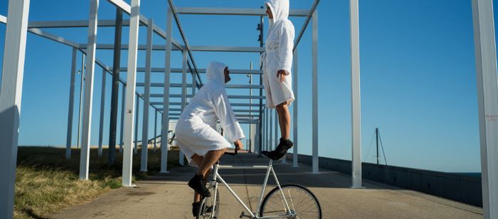 Photographie prise en extérieur sous un ciel bleu dégagé. Au centre de l’image, deux personnes vêtues de peignoirs blancs à capuche et de chaussures noires interagissent avec un vélo blanc.
Une personne est accroupie sur les pédales du vélo, maintenant l’équilibre. L’autre personne se tient debout sur le guidon et semble légèrement surélevée au-dessus du vélo. L’ensemble évoque une figure d’équilibre ou une acrobatie.
La scène se déroule sur une allée en béton bordée d’herbe. Autour des…