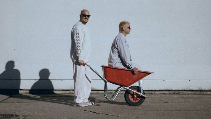Photo des deux membres du groupe dont l'un pousse une bouette avec l'autre à l'intérieur. Ils sont habillés en blanc et portent des lunettes de soleil
