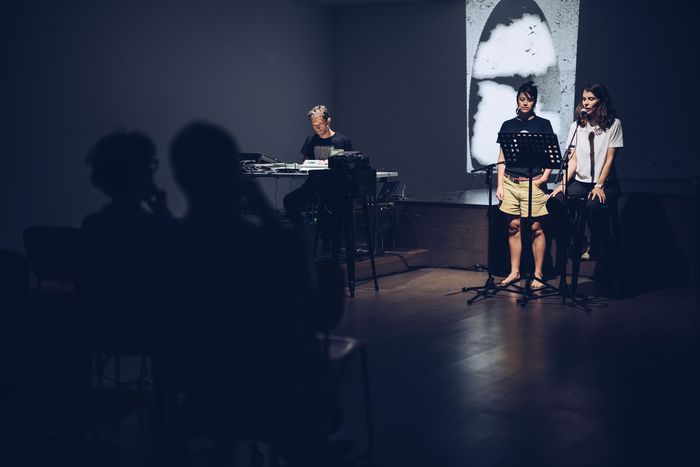 Photo d'une lecture sonore avec un musicien assis avec appareils électroniques et deux femmes debout lisant à un micro.