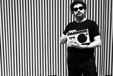 Une photo en noir et blanc : fond rayé et Mr Hobbs avec des lunettes noires et un lecteur de cassettes des années 80.