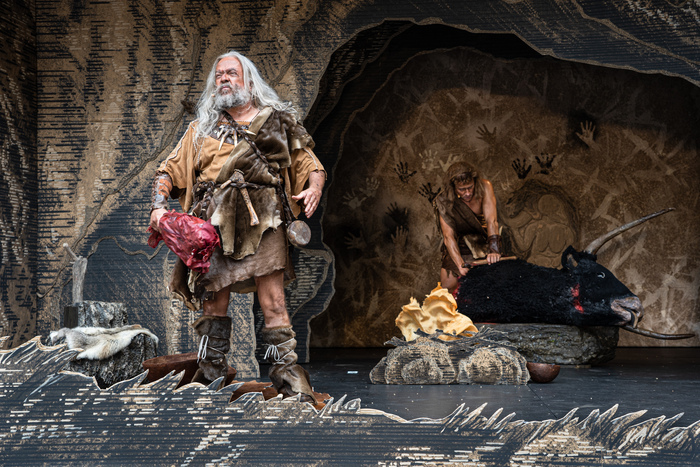 © Théâtre de Carouge / Cédric Vincensini Un homme préhistorique à l'air hargneux, un gros morceau de viande saignante à la main, regarde devant lui, pendant qu'un autre homme préhistorique, derrière, lui dépèce un bœuf mort.