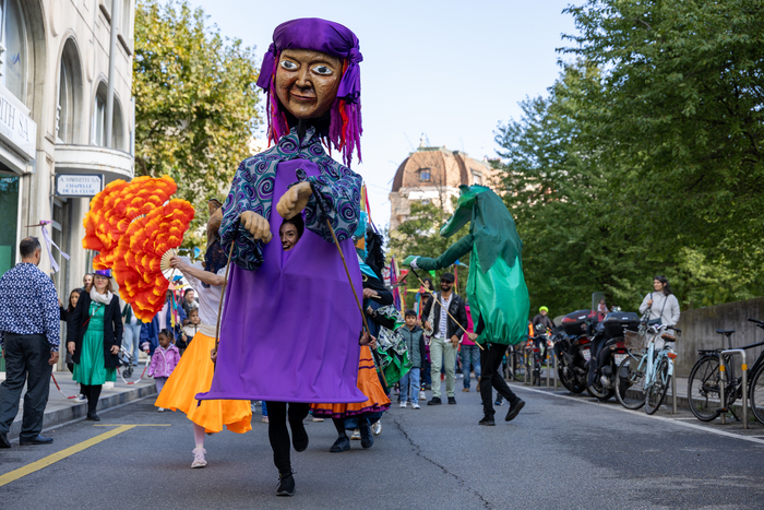 Grande marionnette du personnage zuza dans la rue avec des acteurs et des actrices qui la suivent et des personnes du public autour.