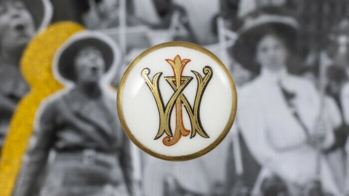 Un bouton en céramique au fond blanc avec des lettres dorés et à la bordure dorée est disposé sur une image en noir et blanc et légèrement floue représentant des femmes.