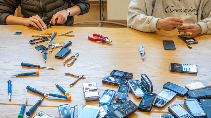 Assis à une table, des personnes dont on ne voit pas la tête sont en train de démonter des téléphones. Sur la table s'étalent des téléphones de toutes sortes, des tournevis et des pinces