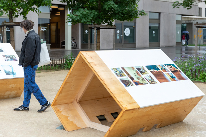Vue d'exposition en extérieur montrant des supports en bois arborant des oeuvres photographiques, ainsi qu'un jeune visiteur.
