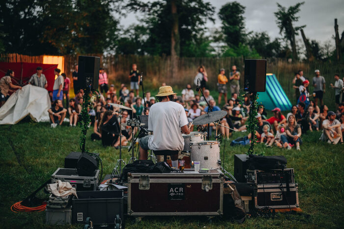 L'image met l'accent sur un batteur de dos. Il fait face à une foule attentive assise dans l'herbe. Le matériel de sonorisation entoure l'artiste, créant une scène improvisée au cœur de la nature.