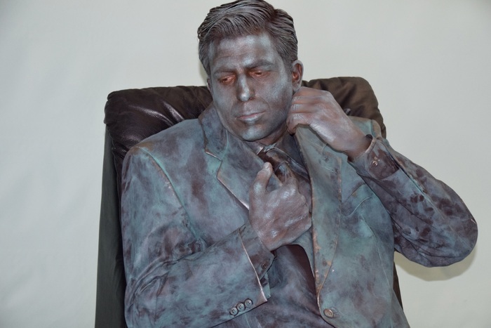 Rebecca Cosne Homme habillé en costume avec son corps et vêtements peints en gris argenté. Il fait face à la caméra et est assis sur une chaise en cuir devant un mur gris clair. Son regard est au sol à droite alors que sa main gauche tient le col du costume prêt de son cou et sa main droite tient la partie proche de sa poitrine.