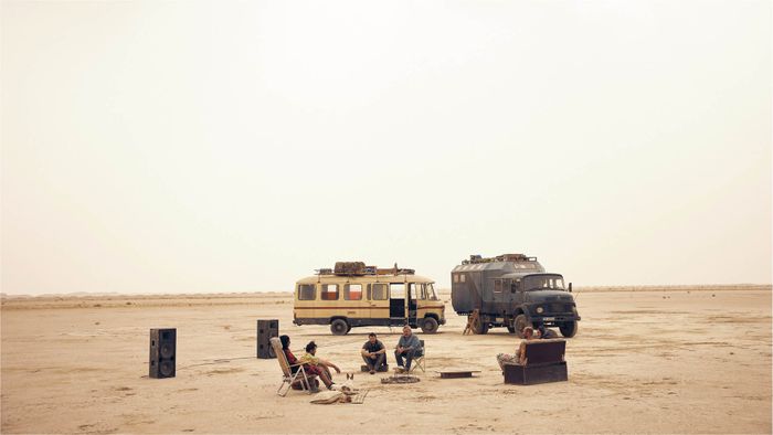 Un desert, deux voitures et plusieurs personnes assises sur des chaises et un canapé devant les voitures. Il y a également deux grosses enceintes pour du son.
