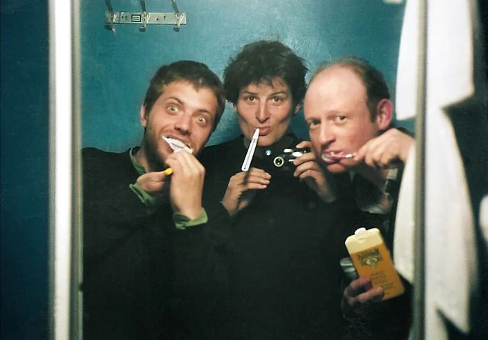 Trois personnes se prennent en photo dans un miroir tout en se brossant les dents, souriantes et décontractées, dans un espace exigu à l’éclairage tamisé.