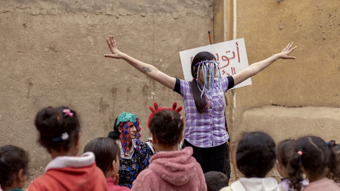 © Les Filles du Nil Performance de rue animée par deux femmes costumées devant un groupe de jeunes filles, dans une ruelle. L’une lève les bras, l’autre est assise. Une pancarte en arabe est visible en arrière-plan. Ambiance ludique et engagée.