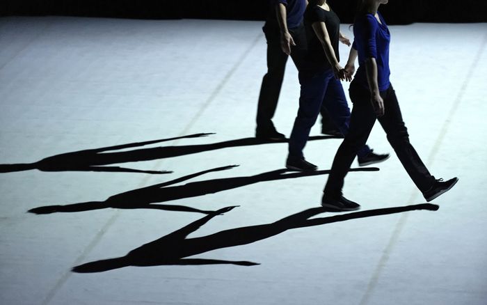 Trois danseurs marchent en ligne, avec un focus sur leurs ombres.