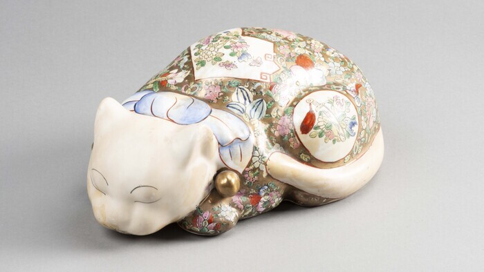 Figurine de chat en porcelaine qui provient de Chine et qui est daté du 20e siècle. Il est allongé, les yeux fermés. Sur son dos se trouve de jolis décors de fleurs.