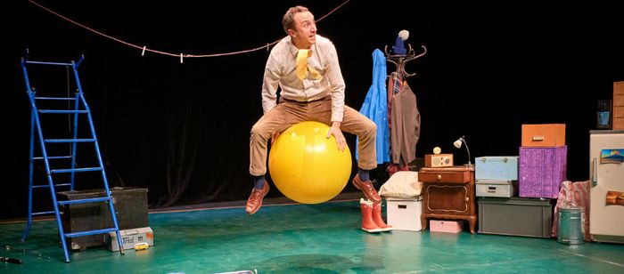 Une scène de théâtre est aménagée avec de nombreux objets du quotidien. Au centre, une personne est assise à califourchon sur un grand ballon jaune de gymnastique. La personne porte une chemise claire, un pantalon beige et des chaussures marron.
À gauche de la scène se trouve une échelle bleue. Un fil à linge est tendu en hauteur d’un côté à l’autre de l’espace, avec plusieurs pinces à linge accrochées. Derrière la personne, un porte-manteau supporte plusieurs vêtements colorés.
À droite, un…