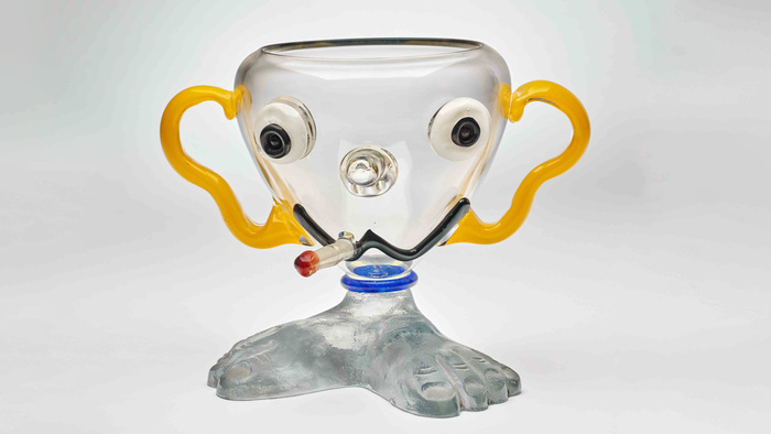 Œuvre de Buddy Di Rosa. Il s'agit d'une coupe en verre transparent sur lequel un visage a été créé en volume, avec une cigarette à la bouche.