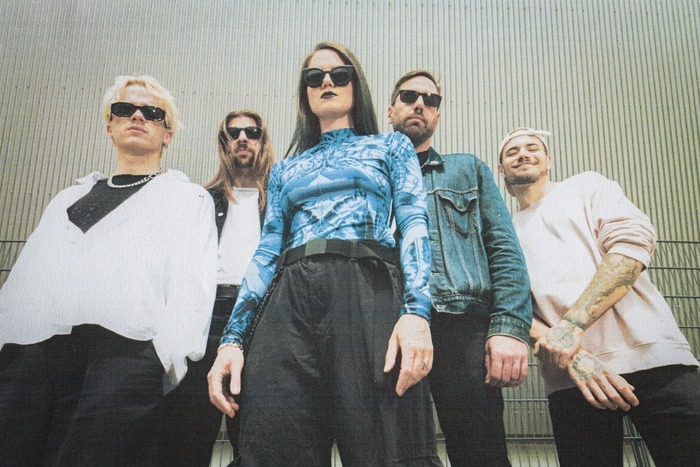 Kassogtha Photo promotionnelle du groupe suisse de metal progressif Kassogtha. Les cinq membres posent devant un mur métallique, regardant la caméra avec assurance. La chanteuse, au centre, porte un haut bleu vif et des lunettes de soleil. Les autres membres, habillés de façon décontractée, portent également des lunettes noires. L’ambiance générale est sombre et puissante.