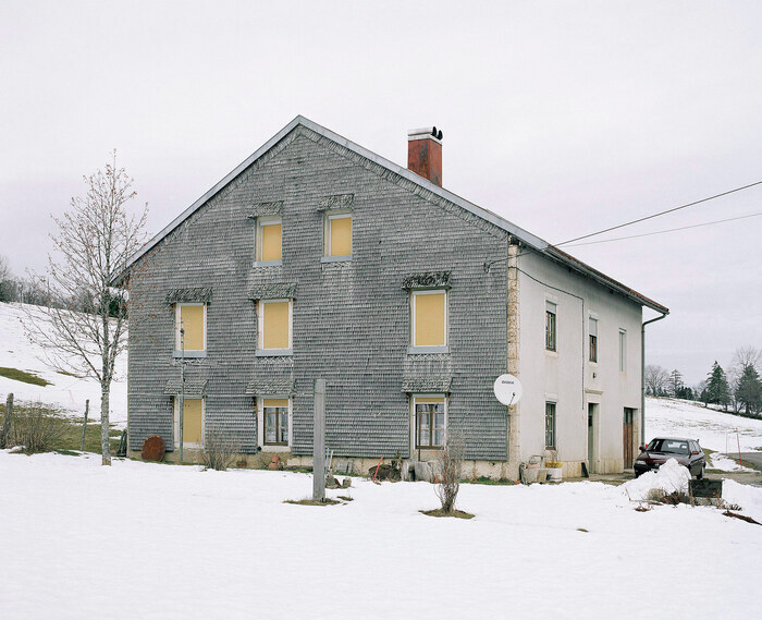 Photographie d’une maison jurassienne grise au volets jaunes fermées, dans la neige.