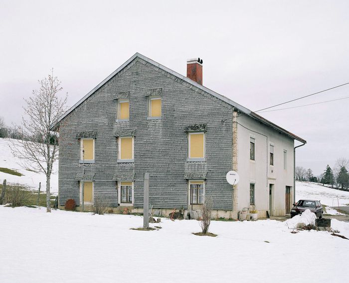 Photographie d’une maison jurassienne grise au volets jaunes fermées, dans la neige.