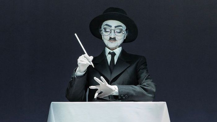 Une personne habillée en mime est assis à une table avec une nappe blanche sur fond noir. Elle porte un costume noir, des gants blancs et une cravate noire.