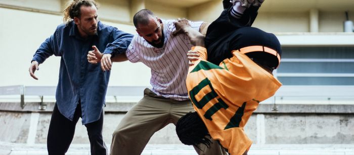 Photographie prise dans un espace extérieur ou semi-ouvert aux couleurs claires. Trois personnes occupent le centre de l’image et semblent participer à une performance de danse ou de mouvement.
À gauche, une personne porte une longue chemise bleu foncé et un pantalon sombre. Les bras sont pliés et légèrement écartés du corps dans une posture dynamique.
Au centre, une personne vêtue d’une chemise claire à fines rayures verticales et d’un pantalon beige est penchée vers l’avant. Un bras est posé…
