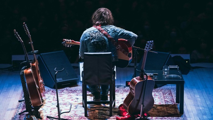 Photo de dos de Ryan Adams sur scène avec plusieurs guitares acoustiques autour de lui