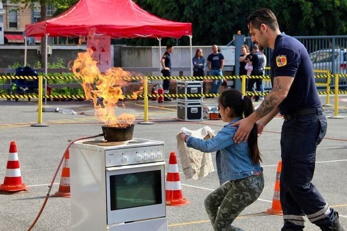 Lors d'une démonstration de sécurité sur un parking, une enfant, guidée par un sapeur-pompier, s'approche d'une casserole en flammes sur une cuisinière, un linge à la main pour étouffer le feu.