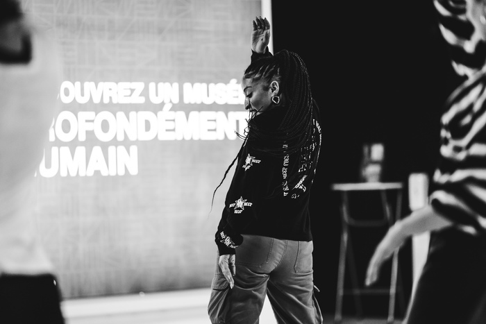 Photo d'un cours de danse au MEG. La professeurs et de côté et les participant-e-s de dos. Il y a des enfants et des adultes qui participent.