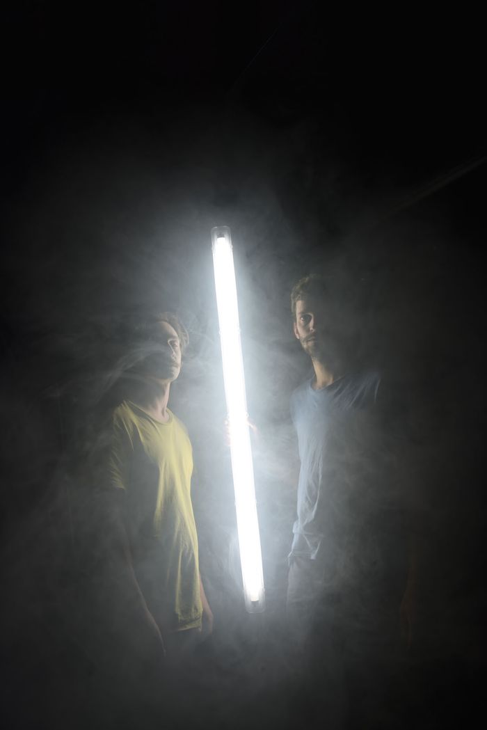 Deux comédiens sont dans le noir, éclairés uniquement par un tube lumineux placé entre eux.