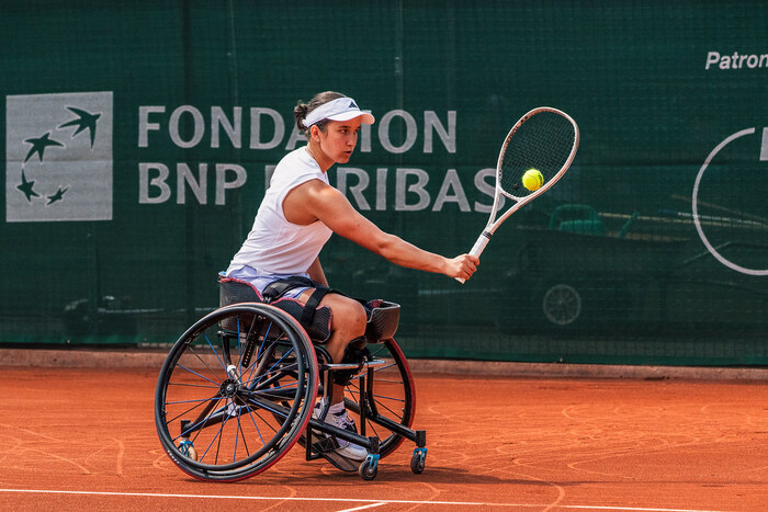 On voit sur cette image, une joueuse de tennis en fauteuil roulant en pleine action sur un court en terre battue.