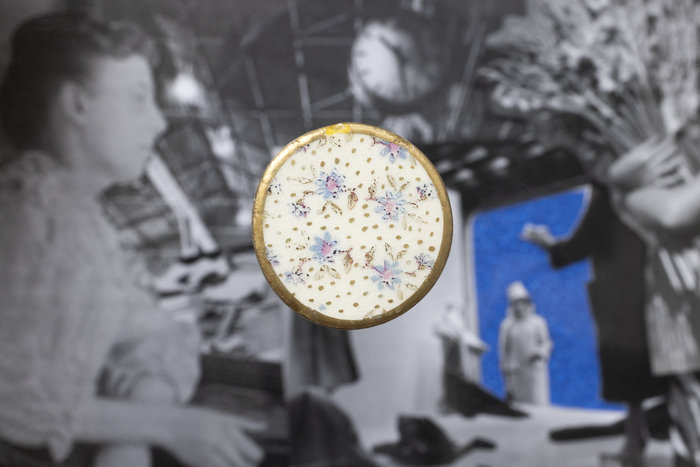Un bouton en céramique au fond blanc avec des décors floraux et à la bordure dorée est disposé sur une image en noir et blanc et légèrement floue représentant des femmes.
