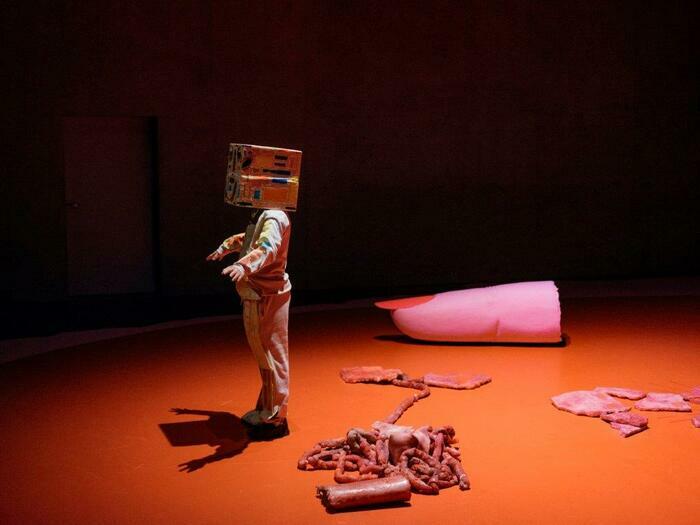 un interprète debout un carton en guise de masque. Des éléments de décors au sol: un doigt coupé, des viscères