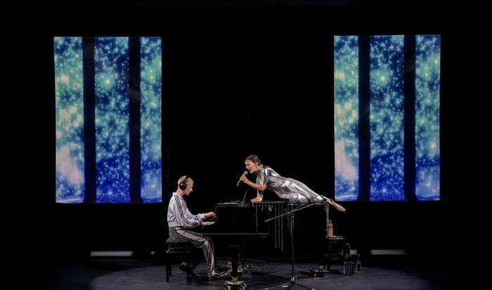 Un homme joue du piano, un casque sur les oreilles. Devant lui, une femme est allongée, un micro à la main. Tous deux portent des vêtements argentés. Deux panneaux lumineux sont placés de chaque côté de la scène