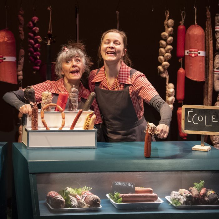 Une photo en couleur sur laquelle on peut voir deux comédiennes, en tablier et chemise à carreaux, dans un décors de fausse boucherie avec des différentes sortes de saucisses sur l'étalage et suspendues.