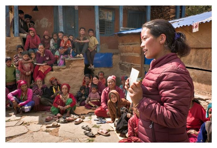 Une séance de sensibilisation de l’organisation partenaire Sahakarmi au Népal en 2025. On voit, de côté, une femme népalaise qui tient une fiche dans ses mains. Elle est en train de parler à une assemblée communautaire dont on voit quelques membres, femmes, hommes et enfants, assis dans un cercle autour d'elle au sol et sur des escaliers d'un bâtiment au fond de l'image.
