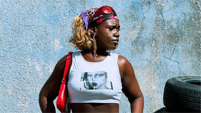 Une femme avec un tshirt représentant une femme avec une sucette pose devant un mur bleuté et des pneus sur la droite de l'image.