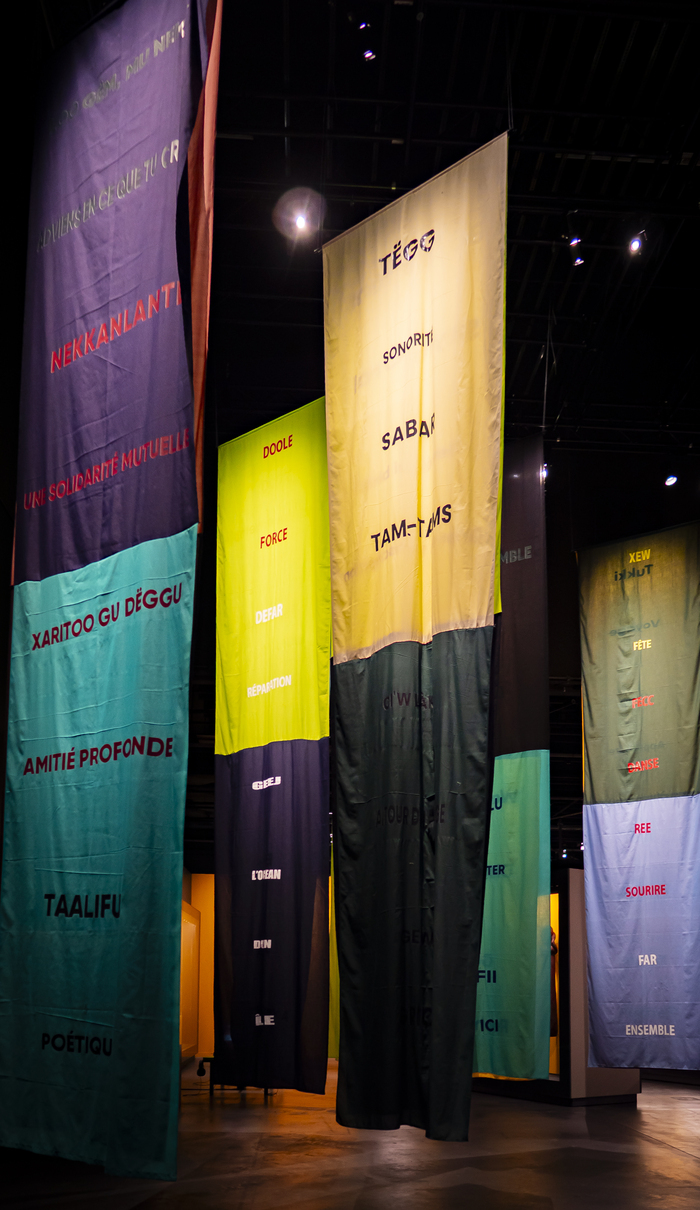 © MEG Oeuvre faite de long tissus coloré qui descendent au sol. L'artiste, Bocar Niang, évoque des mots, des griots modernes. Les mots sont en français et en wolof.