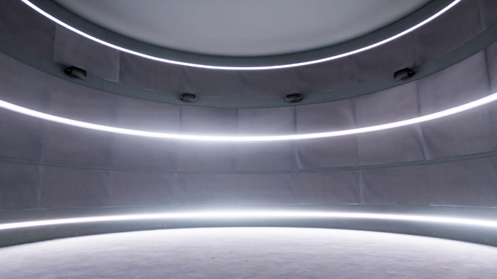 Espace dans l'exposition Afrosonica. Vision panoramique d'un espace circulaire sombre. Trois lignes circulaires de néon, du sol au plafond structure le tout. L'image est dans les ton sombres et gris et les néons ressortent très lumineux.