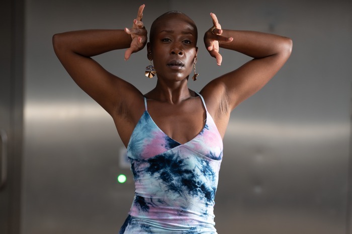 Photo de Laëtycia Vumuka de face dans une posture de danse. Une jambe levée et les deux bras replié vers son visage qui nous regarde. Elle a une robe avec du bleu, rose et blanc et des boucle d'oreille dorées.