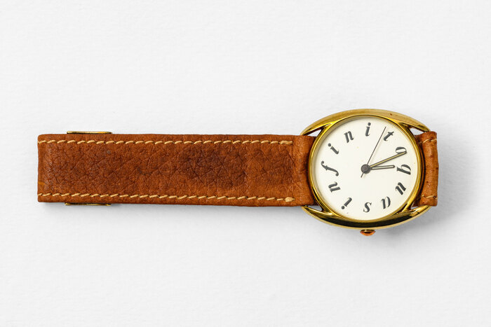 Montre bracelet au cadran doré et bracelet en cuir, avec le mot "quasinfito" en lettres italiques remplaçant les chiffres tout autour du cadran.