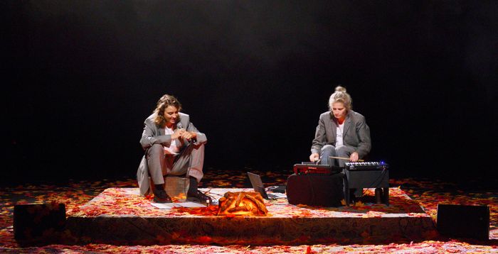 Deux comédiennes sont assises sur scène. L'une avec un micro dans les mains, l'autre bat le rythme avec des baguettes. Le sol est recouvert de feuilles mortes rouges et oranges.