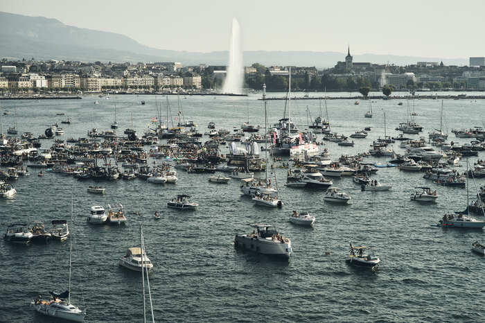 Une foule impressionnante de bateaux est rassemblée sur le lac Léman lors de l'événement On The Water Geneva Festival. En arrière-plan, on observe le Jet d'eau de Genève, avec une vue sur la ville, la cathédrale et un aperçu du Salève