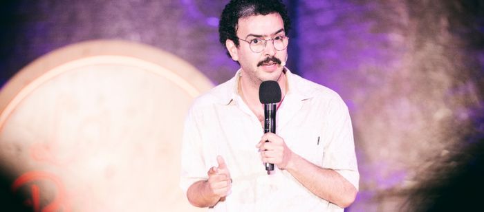 Une personne se tient sur une scène et parle dans un microphone tenu à la main. La personne porte des lunettes rondes, une chemise claire à manches courtes et un micro-casque fixé près de la joue. Une moustache est visible.
Le cadrage est serré sur le haut du corps. Une main tient le microphone tandis que l’autre est légèrement levée dans un geste d’accompagnement du discours.
L’arrière-plan est flou et éclairé par des lumières violettes et rosées. À gauche de l’image, on distingue une grande…