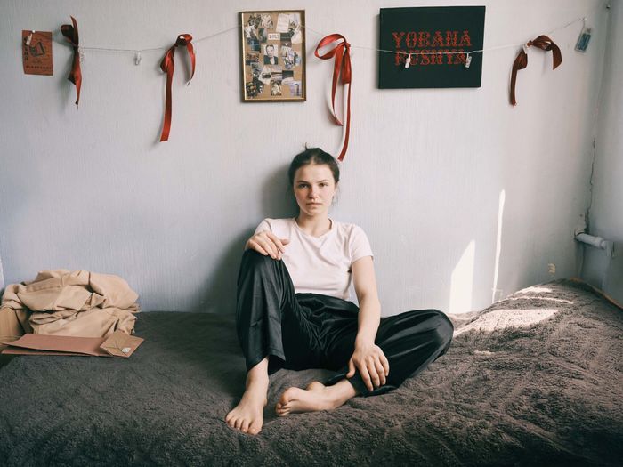 Une jeune fille est assise sur son lit, appuyée contre le mur décoré de sa chambre. Un cadre, rempli de photographies personnelles avec la mention 2025, un panneau où y est écrit en lettres rouges Yobana Pusnya (en ukrainien) et des rubans rouges sont suspendus au mur.