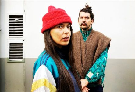 Deux personnes, une femme et un homme, font face à la caméra et portes des vêtements colorés - la femme porte un bonnet rouge. Ils sont face à un mur plutôt industriel / urbain, avec des bouches d'aération et un tuyau de descente (gouttière).