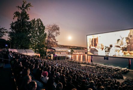 PPR Media Relations AG Un grand écran de cinéma en plein air est installé au bord du lac Léman, avec une foule de spectateurs assis en gradins. La projection a lieu à la tombée de la nuit, sous un ciel violet orangé avec la lune brillante. En arrière-plan, on aperçoit le lac, des arbres et un bâtiment éclairé. L’ambiance est calme et magique.