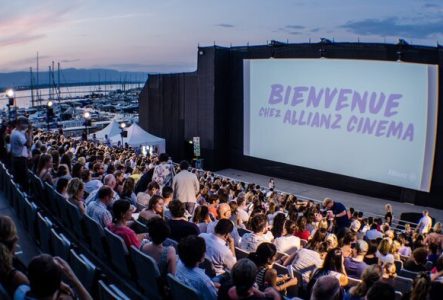 Un grand écran de cinéma en plein air est installé au bord du lac Léman, avec une foule de spectateurs assis en gradins. La projection a lieu à la tombée de la nuit, sous un ciel violet orangé avec la lune brillante. En arrière-plan, on aperçoit le lac, des arbres et un bâtiment éclairé.