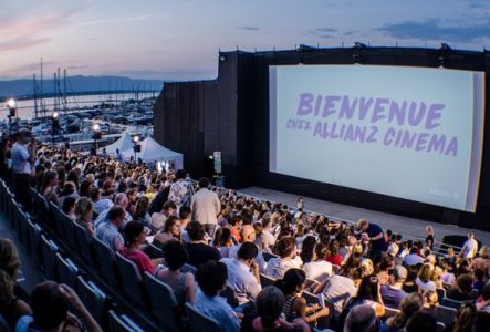 Un grand écran de cinéma en plein air est installé au bord du lac Léman, avec une foule de spectateurs assis en gradins. La projection a lieu à la tombée de la nuit, sous un ciel violet orangé avec la lune brillante. En arrière-plan, on aperçoit le lac, des arbres et un bâtiment éclairé.
