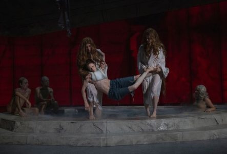 Une piscine en pierre est sur scène. Deux danseurs sortent de l'eau une femme inconsciente. Trois autres sont assis, à l'arrière-plan.