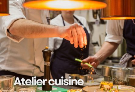 Photographie en gros plan dans une cuisine professionnelle montrant un chef de profil, en veste blanche, qui saupoudre d'épices une assiette soigneusement dressée. En arrière-plan, d'autres cuisiniers en tablier bleu s'activent sous des lampes chauffantes en cuivre. En haut à gauche figure le logo blanc de l'Alliance Française Genève - Suisse. En bas, le texte en blanc indique : "Atelier cuisine - Initiation à la Gastronomie créole".