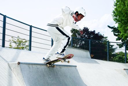 ©Rookie Slash Un jeune skateur habillé en blanc. Il porte un casque blanc et effectue une figure sur une rampe en béton dans un skatepark en plein air.