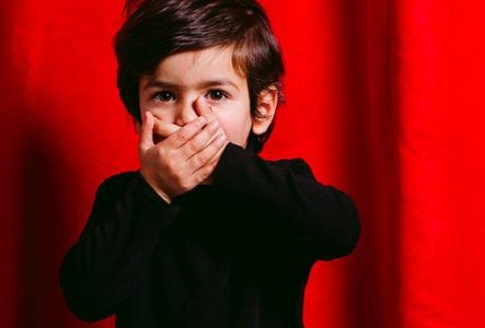 Un enfant se met les mains devant la bouche l'air surpris. Il se tient devant un rideau de théâtre rouge fermé